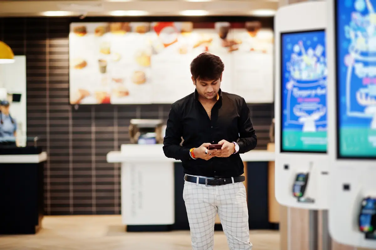 Stylish indian man posed at fast food cafe near self pay floor kiosk with mobile phone at hand